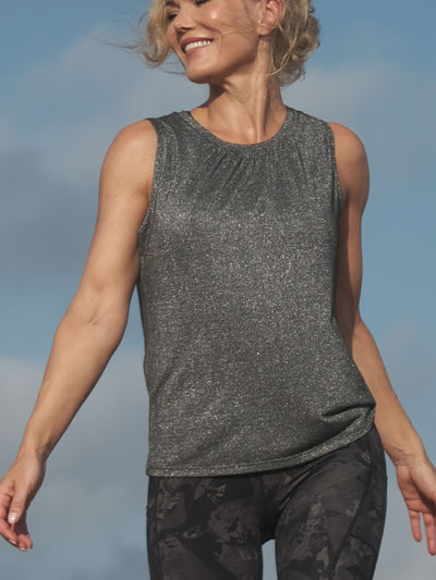 Kvinne som smiler utendørs under blå himmel i glitrende, mørkegrå tank topp med flatterende snitt og fine detaljer, stylet sporty med svarte treningstights med kult design.