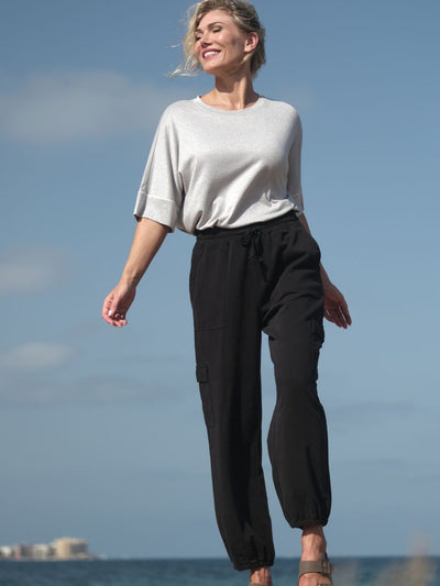 Kvinne ved sjøen i sølvglitrende T-skjorte med myk passform og halvlange ermer, stylet med svart joggebukse for en avslappet look.