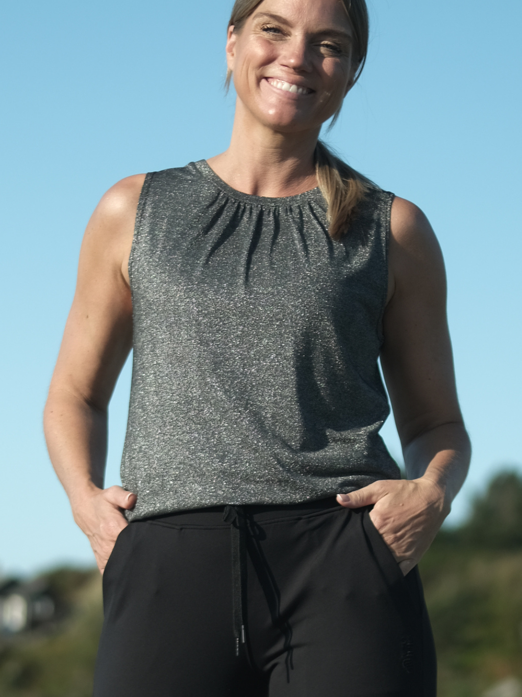 Kvinne som smiler og poserer med hender i lommene utendørs i glitrende, mørkegrå tank topp med flatterende snitt og fine detaljer, stylet sporty med svarte joggebukser.